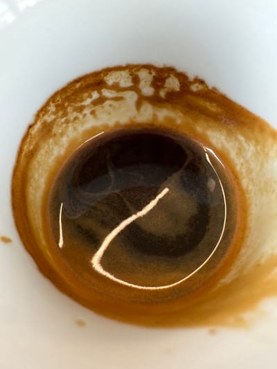A close-up, top-down view depicts the interior of a white ceramic cup, located in Anzola dell'Emilia, Italy. The cup contains a small volume of dark, reddish-brown liquid at its base, consistent with a coffee beverage such as espresso. Surrounding the liquid is a prominent, light-brown ring of crema, which exhibits an irregular, textured, and slightly foamy pattern adhering to the inner wall of the cup. Two distinct, bright, curved reflections are visible on the surface of the liquid: one longer, arcing diagonally across the lower-center, and a shorter, vertical reflection positioned above and to the left of the liquid's center. The white ceramic of the cup is visible as a clean background around the liquid and crema. No individuals, specific actions, or other objects are visible within the frame. The scene is static, captured under diffused lighting conditions.