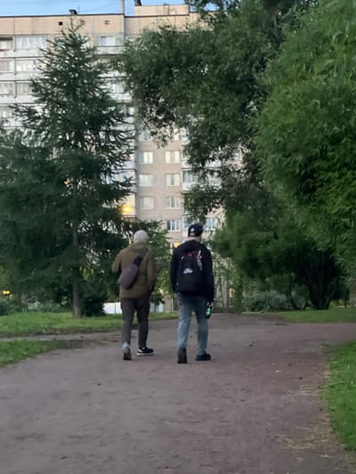 Two people are walking along a dirt path next to a line of trees and a building. The person in the foreground is wearing a light brown jacket, dark pants, and a white cap. They have a light purple bag slung across their body. The second person is behind and to the left of the first, wearing a dark jacket with patches on the back, and blue jeans. They are wearing a black helmet and appear to be holding a green object. The building behind the trees is a multi-story apartment complex with numerous windows. The scene appears to be in a park or residential area with green grass and trees. The lighting suggests it is either early morning or late evening, with a soft, diffused light. No text is visible in the image.