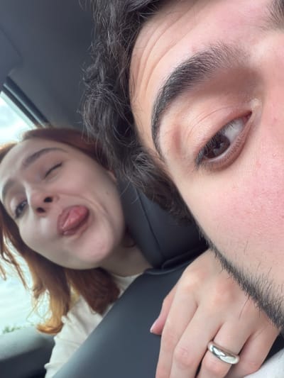 Two individuals are in what appears to be a vehicle. One person is positioned in the upper part of the frame with a close-up view of their eye, eyebrow, and part of their beard. A ring is visible on their finger. The other person is in the lower part of the frame, sticking out their tongue and winking. The environment appears to be inside a car. There is no verifiable information in the image to determine whether the picture was taken in Tserovani, Georgia.