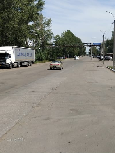 The image shows a street scene in Aleysk, Russia, with a white semi-trailer truck on the left side of the road. The truck has Chinese characters on its side. In the middle of the road, a beige sedan is driving away from the camera. The road is paved and appears to be in moderate condition with some patches. Trees line both sides of the road, and a blue sign with white lettering, "АВТОВЕСОВАЯ" (Autoweighing Station), is visible overhead on a metal structure. Several other vehicles are parked along the right side of the road. The sky is partly cloudy. The watermark "Galaxy A05s" is present in the bottom left corner.