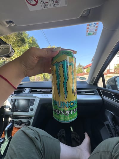 In the city of Zaprešić, Croatia, a person's hand holds a yellow and green can of Monster Energy drink, identified as "Punch Monster," inside a car. The car's interior is visible, with the dashboard featuring a media console and climate controls. Outside the car, through the windshield, a residential area with houses and trees can be seen under a clear blue sky. The person is wearing green pants and is barefoot, with one foot resting on the car's floor. A red string bracelet is worn on the wrist holding the can. A sticker is attached to the windshield.