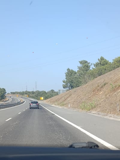 The image shows a view from inside a car traveling on a highway. The highway curves to the left and has multiple lanes, separated by white lines. A gray car is in front of the camera's car, also on the highway. On the right side of the highway, there is a steep, dry, grassy embankment with some scattered green plants. Tall pine trees and other vegetation grow at the top of the embankment. On the left side of the highway, there are more trees and a concrete barrier with metal fencing. In the distance, power lines and transmission towers are visible against a clear blue sky. Several small, round, colored objects, possibly balloons, are scattered in the sky. The scene appears to be in a rural area with arid vegetation, consistent with the landscape around Santiago do Cacém, Portugal, particularly along its highway routes.