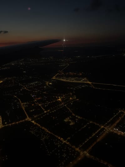 The image is an aerial view of a city at dusk, likely taken from an airplane. The foreground is dominated by the dark silhouette of an airplane wing. Below, the city lights twinkle through the fading daylight, creating a network of illuminated streets and buildings. The sky transitions from a deep blue at the top to a band of orange and red near the horizon. A bright light, possibly a star or the moon, is visible in the upper portion of the sky, near the plane's wing. The overall impression is one of a city settling in for the night, viewed from above. The location in the city of Шемякино, Russia is not discernible from the image. There are no people, specific subjects, actions, or interactions visible.