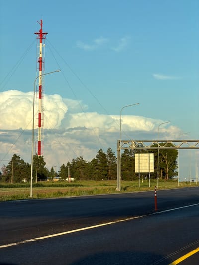 A tall, red and white striped radio tower stands prominently on the left side of the frame, its cables extending outwards and disappearing into the sky. A street lamp is positioned in front of the tower. The sky above is a brilliant blue, dotted with large, puffy white clouds. In the middle ground, a dense line of evergreen trees borders a field of green grass and crops. To the right, a highway-like road stretches into the distance, with multiple street lamps lining its edge. A metal gantry with a blank white sign hangs over the road. A single orange traffic cone stands on the shoulder of the road in the foreground. The overall scene appears to be located in a rural or semi-rural area, potentially on the outskirts of Egor'evo, Russia, given the presence of the radio tower, fields, and highway infrastructure.
