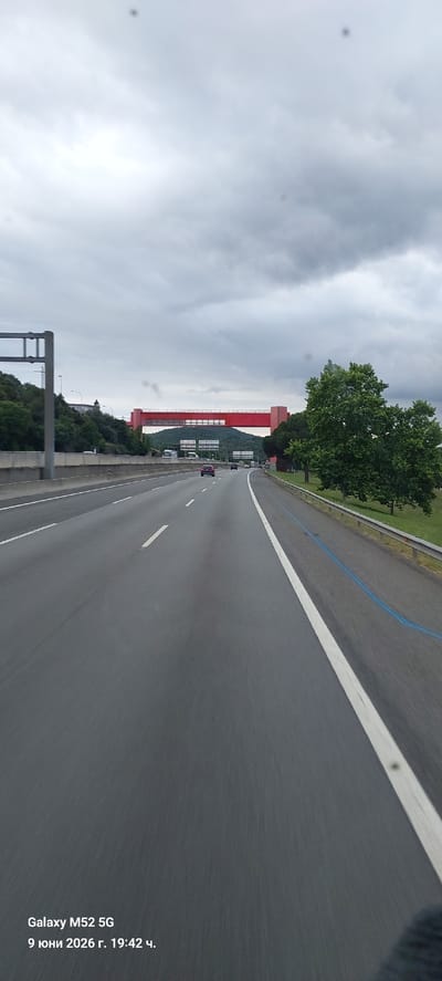 The image shows a view from a vehicle traveling on a highway. A bright red overpass stretches across the highway in the middle distance, with a green, tree-covered hill behind it. The sky is overcast and gray. On the right side of the highway, there is a guardrail and a grassy embankment with trees. A blue line is visible on the right shoulder of the road. The bottom left corner of the image displays a watermark indicating "Galaxy M52 5G" and the date and time "9 юни 2026 г. 19:42 ч." (June 9, 2026, 19:42). The scene is likely located on a highway near Maçanet de la Selva, Spain, given the date format which suggests a localized setting. There are no people clearly visible, and the primary action is vehicular movement on the highway.