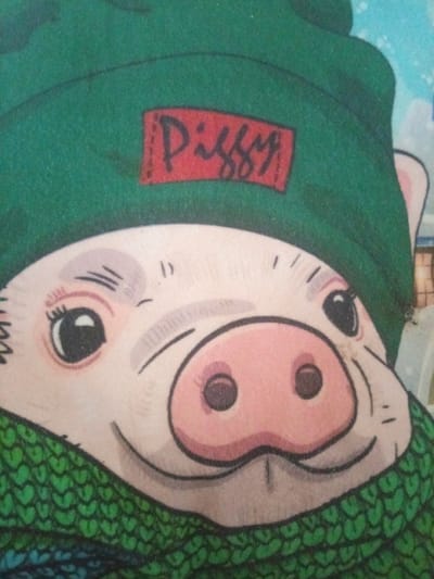 The image is a close-up, stylized illustration of a pig's face wearing a green hat and a green scarf. The pig has large, dark eyes with eyelashes, a pink snout with two dark nostrils, and a smiling mouth. Above its eyes, there are subtle greyish blush marks. The green hat has a red rectangular patch on the front with the word "Piggy" written in black, stylized cursive lettering. The green scarf is knitted with a pattern resembling small hearts. The background is out of focus, showing a light blue sky with white splotches that could be clouds or snow. The overall impression is that of a character from a children's book or animation, conveying a cheerful and friendly emotion. There are no indications of the time of day, weather conditions, or a specific location beyond what is depicted in the illustration itself.