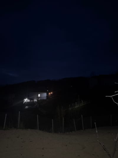 This image depicts a nighttime scene in the city of Byal Izvor, Bulgaria. The central focus is a two-story, light-colored residential structure situated on a hillside. Multiple windows and exterior lights on the house are illuminated, casting a warm glow onto its immediate surroundings and a portion of a gravel or dirt driveway where a parked white vehicle is partially visible.

The house is partially obscured on its left by a darker, possibly overgrown area, while a small, dark wooden shed or outbuilding with an illuminated exterior light is visible to its right, further up the slope. The terrain around the house is uneven and vegetated with sparse, bare trees and shrubs.

In the foreground, a wire fence with wooden posts runs horizontally across the lower third of the frame, separating a dark, earthy ground area from the illuminated house and hillside beyond. The upper portion of the image is dominated by a dark blue-black night sky, with a subtle lighter band visible on the far left horizon, indicating very early morning or late evening twilight. No individuals or specific actions are discernible within the frame.