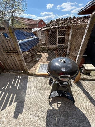 The image shows a backyard scene in Gabarevo, Bulgaria. A black charcoal grill is positioned on a concrete surface next to a dilapidated wooden fence topped with chain-link fencing. Behind the fence, there's an old, weathered shed-like structure with a tile roof and a fenced-in window. A blue tarp is partially visible behind the fence. In the background, sections of neighboring buildings with red tile roofs can be seen under a blue sky with scattered clouds. The sun is shining, casting shadows from the fence and the grill onto the concrete.