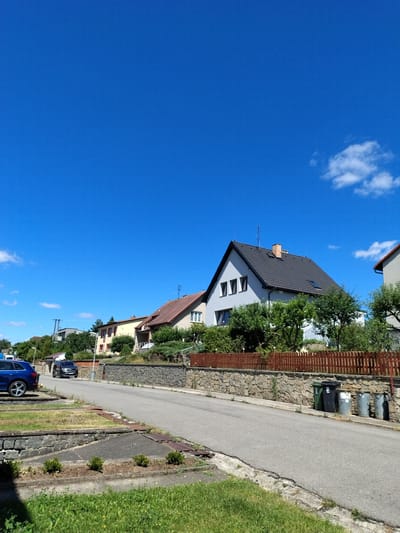 In the city of Dobev, Czechia, a residential street is depicted under a clear blue sky with scattered clouds. The street is paved and curves slightly to the right, bordered by a stone wall on the left and a wooden fence on the right. Several houses with pitched roofs are visible on the hillside behind the fence. In the foreground, on the left side of the street, a blue SUV is parked. Further down the street, a black car is in motion. Several trash bins are placed near the stone wall on the right side of the road. Green grass and small bushes grow along the edges of the street and on the embankment.