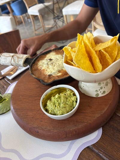 A hand is reaching towards a cast iron skillet filled with a cheesy, bean-based dish, accompanied by tortilla chips in a decorative bowl and a small dish of guacamole. The scene appears to be set on a wooden board, with a white placemat underneath. In the background, several chairs with blue and white upholstery are visible, suggesting a restaurant or cafe setting in Rionegro, Colombia. The lighting indicates an indoor environment, possibly during the daytime.