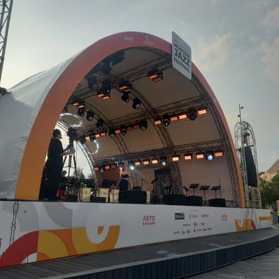 This image captures an outdoor stage set up for the Moscow Jazz Festival. The stage has a large, curved white and orange awning supported by a metal truss structure. Numerous spotlights and lighting rigs are mounted on the truss, with some illuminated. A singer, dressed in black, is on stage performing into a microphone. To the left of the singer, a person in black is operating a professional video camera on a tripod. In the background, musical instruments and music stands are visible, suggesting a band or ensemble is present. The front of the stage is adorned with a white banner featuring colorful geometric patterns and logos, including "ЛЕТО В МОСКВЕ" (Summer in Moscow) and various sponsor names. The setting appears to be daytime with a bright, slightly overcast sky. The scene is taking place in a public outdoor area, likely a plaza or park. The image does not provide any information about the specific location within the city of Путрышкаўскі сельскі Савет, Belarus.