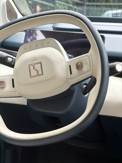 The image shows a close-up of the steering wheel and dashboard of a car. The steering wheel is two-toned, with a cream-colored leather grip and a dark grey outer rim. A gold emblem, possibly a car logo, is visible in the center of the airbag cover. The dashboard features a screen and several buttons with icons, including what appears to be a heated steering wheel symbol, a camera symbol, and a driver assistance symbol. A gear shifter with a "P" for park is visible on the right side of the steering column. The interior of the car appears to be modern and clean, with a dark blue trim accenting the cream and grey. The background shows a blurry view of what appears to be a street with trees, suggesting the car is parked outdoors. There is no indication of the city of Mozhaisk, Russia in the image.