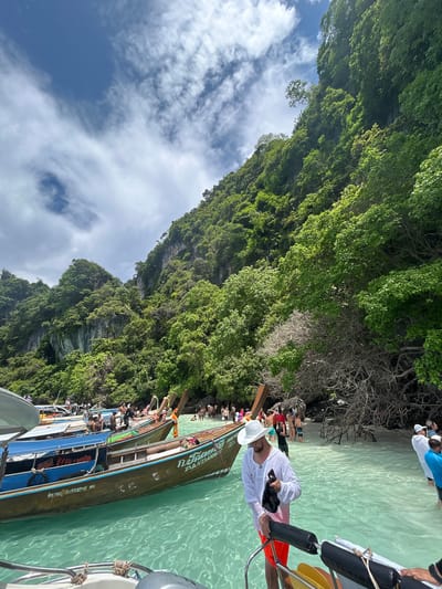 The image depicts a sunny day at a beach in Ko, Thailand, likely on Phi Phi Island, known for its limestone cliffs and clear turquoise waters. In the foreground, a man wearing a white hat and white long-sleeved shirt over orange swim shorts stands on the edge of a boat, holding a black bag. The boat is a traditional longtail boat, with Thai writing visible on its side. Several other longtail boats are moored in the shallow, crystal-clear water.
In the background, a lush green, heavily forested cliff face rises steeply from the water. The beach is populated by numerous people, some wading in the water, others standing near the boats or walking along the shore. The sky is partly cloudy with bright white clouds against a blue backdrop. The overall scene conveys a vibrant tropical tourist destination.
