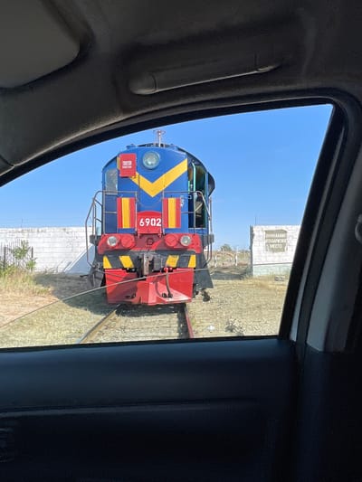 A blue and yellow diesel locomotive with the number 6902 is visible on railroad tracks. The locomotive has red and black accents. A red sign on the front of the locomotive reads "TOKTON". A white brick wall is behind the train to the left, and to the right, a white sign with black lettering that reads "ВНИМАНИЕ! НЕГАБАРИТНОЕ МЕСТО" (Warning! Oversized place). The scene is viewed from inside a vehicle through a car window. The environment appears to be dry and grassy, with a clear blue sky overhead. This image was taken in Ömirzaq, Kazakhstan.
