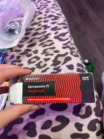 A person's hand is holding a box of "Citramon P" medication manufactured by "Medisorb". The box is predominantly black with red diagonal lines and white text. The text "200 таблеток" meaning "200 tablets" is visible in the upper right corner. The background consists of a leopard-print fabric and a dark surface, possibly a floor or table. The location is stated to be Vaganovo, Russia.