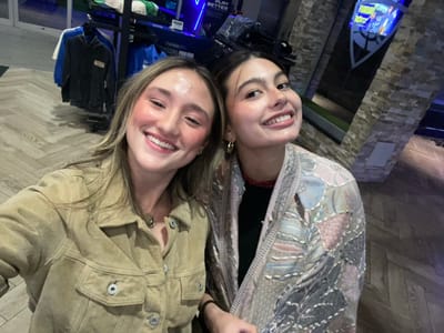 Two smiling young women pose for a selfie, their heads close together. One wears a patterned shawl over a black top, accessorized with hoop earrings. The other is dressed in a tan suede jacket. They stand indoors against a backdrop featuring textured stone paneling and a display rack filled with merchandise, including blue bags and dark-colored apparel. The floor appears to be herringbone-patterned wood. The image has been rotated, positioning the subjects sideways.
