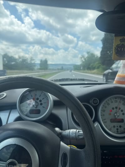 The image captures a first-person perspective from the driver's seat of a Mini Cooper, traveling on a multi-lane highway near Kipra, Bulgaria, under a partially cloudy sky. The dashboard, visible in the foreground, features a gray steering wheel with a "MINI" logo at its base and some visible wear. To the right of the steering wheel, a wiper control stalk is partially visible. The circular tachometer on the left shows the needle between 2 and 3 (approximately 2500 RPM), with a digital display below indicating "23.5". The speedometer on the right indicates a speed just under 120 (likely km/h), and a digital display below reads "123006" with a red warning light illuminated. A partial view of a radio or infotainment screen at the bottom right shows "FM 103.8".
Through the windscreen, a paved highway is visible, extending into the distance with distant vehicles. Trees and dense green foliage line the road on both sides. In the far distance, hills or mountains are discernible under a pale blue sky dotted with numerous white cumulus clouds. A dark-colored sedan is partially visible on the right side of the road, while a large white vehicle or truck is seen in the left distance. A "VIP" branded air freshener, yellow/gold with a crown motif, hangs from the rearview mirror, which is partially visible at the top center of the frame.