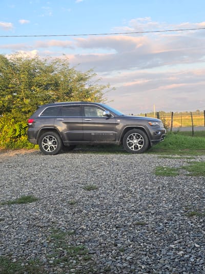 A dark gray Jeep Grand Cherokee is parked on a gravel driveway. The vehicle is facing the right side of the frame, with its rear end slightly closer to the viewer. Lush green trees with vibrant foliage are visible to the left of the Jeep. In the background, a wire fence runs horizontally across the scene, separating a grassy field from a road. The sky above is a soft blue, dotted with wispy white and pink clouds, indicating it is either early morning or late afternoon. The overall environment suggests a rural setting, possibly on the outskirts of Ingiri, Georgia.