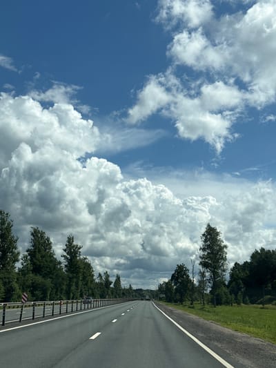 An asphalt road extends into the distance, bordered by a metal guardrail on the left and a grassy verge and trees on the right. The sky above is a bright blue, filled with dramatic, voluminous white clouds. A single vehicle is visible on the road ahead. Red and white striped markers are attached to the guardrail. The scene suggests a rural or suburban setting, possibly the outskirts of Mshaga, Russia, given the natural landscape and absence of dense urban development.