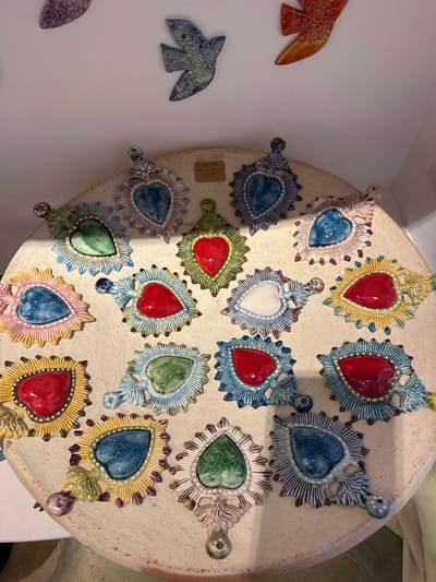 This image depicts a display of handcrafted ceramic items within an artisan shop in Saint-Paul-de-Vence, France. The primary focus is a collection of seventeen colorful, heart-shaped ceramic pendants, designed in the style of sacred hearts with radiating rays, arranged on a circular, light beige display surface. These pendants feature various glossy glazes, including red, multiple shades of blue, green, white, pink, and yellow, often accented with small white dot patterns. A small, light brown rectangular price tag reading "Coûte 25€" is visible among the top center pendants. Above the display, affixed to a plain white wall, are at least three ceramic bird-shaped wall decorations. One blue bird with intricate patterns is centered, flanked by a partial purple bird on the left and an orange-brown patterned bird on the right. No individuals are present in the scene.