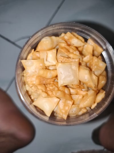 An overhead, close-up shot shows a clear plastic container filled with golden-brown, fried, square-shaped crackers. The crackers are piled high in the container, with many overlapping and some appearing slightly puffed. The container sits on a grey surface with faint white lines, possibly a tiled floor or countertop. The lower left and right edges of the frame show blurry, out-of-focus areas of what appear to be human legs or feet. The overall lighting suggests an indoor setting. The image is taken from a slightly high angle looking down into the container. The location is identified as Barengkrajan, Indonesia.