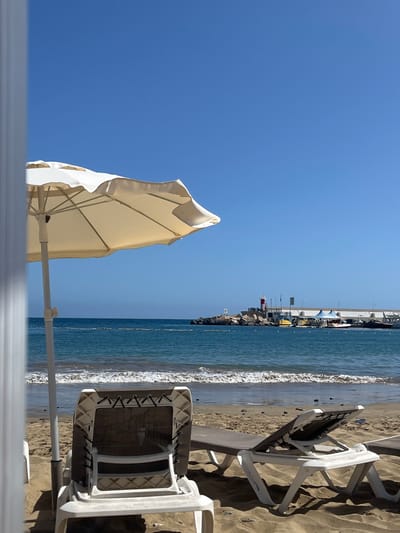 The image shows a beach scene in Puerto Rico, Spain, with a clear blue sky. An off-white beach umbrella is partially visible in the foreground, obscuring a portion of the sky. Adjacent to the sea are two white plastic beach chairs on the sand. The ocean, exhibiting light waves, borders the sand. Further in the distance, a pier-like structure extends into the sea, featuring multiple structures and a red lighthouse-type edifice. The perspective appears to be from an elevated viewpoint. The image is rotated 90 degrees counterclockwise.