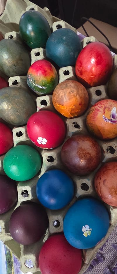 A close-up image captures a collection of numerous brightly colored Easter eggs arranged in a cardboard egg carton, likely in a home in Novachene, Bulgaria. The eggs display a variety of hues, including vibrant reds, blues, and greens, as well as more muted orange-brown, dark purple, and metallic-sheened green tones.

Several eggs feature decorative elements: a light blue egg has a small white and yellow/green flower sticker, and a red egg also has a small white flower. Another orange-brown egg exhibits a unique, possibly abstract or butterfly-like, sticker design in purple. Notably, one variegated green, red, and yellow egg bears visible white Cyrillic text, which reads "Христос" (Christos), a traditional greeting associated with Orthodox Easter celebrations.

The eggs are snugly fitted into the individual compartments of the carton, suggesting either preparations for the holiday or their presentation. In the upper left background, a dark, blurry shape possibly indicates a cable or a part of furniture, while a section of patterned fabric with a purple floral design is visible on the right. The overall scene conveys the festive tradition of dyeing and decorating eggs for Easter.