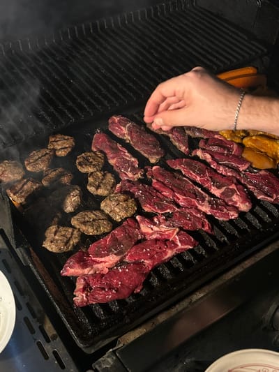 A fair-skinned human hand, wearing a silver chain bracelet on the left wrist, is seen from above, actively sprinkling coarse white crystalline seasoning onto raw, thinly sliced red beef on a black metal grill grate. The grill, located in Yehud Monosson, Israel, is heavily laden with various uncooked and partially cooked food items. Approximately twenty pieces of raw beef, displaying varied shapes and sizes, are arranged across the right and lower-right sections of the grill, with some visible white fat marbling. To the left, eleven round, browned beef patties exhibit distinct char marks, indicating they are in various stages of cooking. In the upper-right corner, two elongated, orange-brown objects, resembling hot dogs or sausages, are visible alongside several pieces of yellowish raw chicken. Wisps of smoke rise from the hot grill surface. Portions of white ceramic plates are visible at the bottom-left and bottom-right edges of the frame.