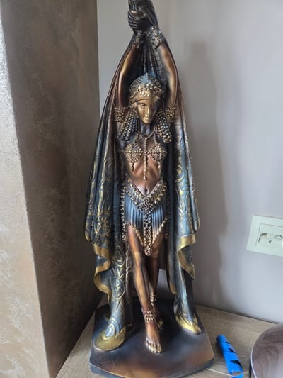 A full-body, vertical, indoor photograph shows a statue of a woman in a regal, ancient Egyptian-inspired outfit. The statue is positioned on a wooden surface against a plain, light-colored wall. The woman is depicted with her arms raised, holding drapery over her head. She wears a golden headdress, a beaded necklace, and a segmented skirt. Intricate patterns and gold detailing adorn her attire and the draped fabric. To the right of the statue, mounted on the wall, is a white electrical outlet with two ports. On the wooden surface, near the base of the statue, lies a blue object, possibly a battery wrapped in tape, and a dark brown, circular object, perhaps a humidifier. The left side of the image features a textured wall with a diagonal shadow. The lighting suggests the presence of a window off-frame to the left, casting a soft illumination. The statue's material appears to be resin or a similar composite, finished with metallic paints. The overall scene suggests a domestic or decorative setting.