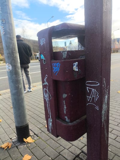 The image displays an outdoor urban scene on a paved sidewalk in Freital, Germany. A weathered, dark red public litter bin occupies the center-right foreground, affixed to a matching dark red vertical pole. The bin and pole exhibit significant wear, including peeling paint, scratches, and multiple graffiti tags in white and blue, along with various stickers. Plastic debris is visible inside the bin's opening. To the left, a gray metal pole, also marked with several stickers, rises vertically from the sidewalk.

The sidewalk consists of gray rectangular pavers, upon which several dry, yellow-orange leaves are scattered near the base of the gray pole. In the midground, a lone individual, viewed from behind and partially obscured by the gray pole, stands or walks on the paved surface. This person is dressed in a black jacket, light grey trousers, and white athletic shoes. An asphalt road with a visible white line runs behind the individual, and a white vehicle is partially discernible in the distance on the road. The background reveals a tree-covered hill, several distant buildings, and streetlights, all set against a blue sky with scattered white clouds.