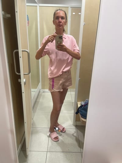 In a changing room, a woman is standing and taking a mirror selfie. She is wearing a pink polo shirt and light pink shorts with frayed edges, and pink slide sandals. The changing room has beige walls and a white tiled floor. The woman is holding a smartphone in her right hand, and her left hand is raised and touching the phone. A dark blue bag with some gray fabric is visible on a wooden shelf in the background. The image is a mirror reflection and shows the woman's front and the interior of the changing room. The scene appears to be in a retail store.
