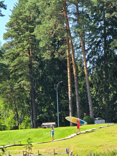 In Voinskaya Chast' Mchs, Russia, a sunny, outdoor scene unfolds with a backdrop of tall pine trees and a clear blue sky. The foreground features a manicured green lawn with several wooden benches and a wooden pathway that ascends a gentle slope.
On the pathway, two individuals are present. One, a person wearing a blue tank top and shorts, walks with a paddle in hand, holding it vertically. Further up the path, another person, dressed in a red t-shirt and red shorts, carries a paddleboard overhead. The paddleboard is yellow and blue.
In the background, more pine trees create a dense forest. A wooden bench is visible on the lawn, accompanied by what appear to be folded towels or blankets. In the far background, stacked white loungers and a light blue mat suggest a recreational area, possibly near a body of water. The lighting indicates a bright, daytime setting.