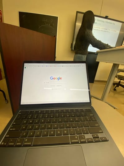 The scene depicts the interior of a classroom or lecture hall located in Lawrence Township, United States. In the immediate foreground, a dark gray MacBook laptop is open and resting on a light-colored desk surface. Its screen displays the Google search engine homepage, primarily in English, with some Hebrew text visible below the search bar.
To the left of the laptop, a dark brown wooden partition or piece of furniture partially obscures a black chalkboard. The visible portion of the chalkboard clearly displays the handwritten text "Oct. 7 Test 3" in white chalk.
In the background, positioned to the right of the wooden structure, a person stands facing a large, illuminated digital display screen. The individual is seen from behind, possessing long dark hair, and is dressed in a dark, possibly knitted, long-sleeved top and light-colored jeans. Their right arm is extended towards the display screen, indicating interaction. The digital screen presents educational content, specifically "1: Solving a System by Substitution (1 of 4)," along with text detailing a mathematical procedure, including "x-coordinate, substitute 3 for x in either original equations and solve for y," and "y = -1."
The wider environment consists of light-colored walls, and a partial view of another table and an office chair on wheels is visible to the far right. The lighting within the room appears to be artificial and warm.