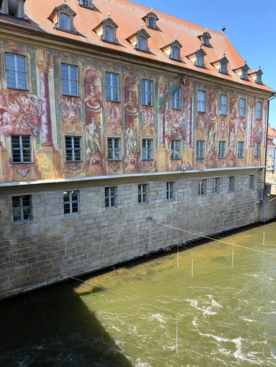 The image shows a section of the Altes Rathaus (Old Town Hall) in Bamberg, Germany. The building is situated on an island in the Regnitz River, and the lower portion of the structure is visible, built of rough stone. Above this stone foundation, the building features a brightly painted facade with elaborate frescoes depicting mythological scenes and figures. A series of regularly spaced windows with blue frames are visible on both the stone and frescoed sections of the building. The Regnitz River flows in front of the building, its surface rippling and showing a greenish-brown hue. Several thin, white vertical poles, some connected by white string, are suspended above the water, creating a grid-like pattern. The sky above is a clear, bright blue.