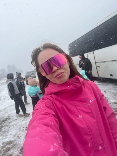 The image shows a person wearing a pink ski jacket and pink-mirrored ski goggles taking a selfie in a snowy environment. In the background, other people are standing and some luggage is visible, suggesting a travel or outdoor setting. A large bus is partially visible on the left side of the frame. Snowflakes are falling. The location is Kliuchi, Russia, judging by the context and other images with similar composition.