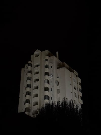 A multi-story building is observed at night in El Campello, Spain. The structure, appearing light-colored under artificial illumination, features a stepped architectural design. Its primary facade includes a vertical column of rounded, glass-enclosed balconies on the left, and recessed rectangular windows on the right. A prominent circular opening is present near the top of the central elevation. The roofline includes a chimney stack. Multiple external air conditioning units are mounted on the facade. A white satellite dish is partially detached from a balcony on the right side. The lower section of the building is obscured by dark, dense tree canopy. The background is a uniform black, consistent with nighttime conditions. No individuals or specific actions are visible.