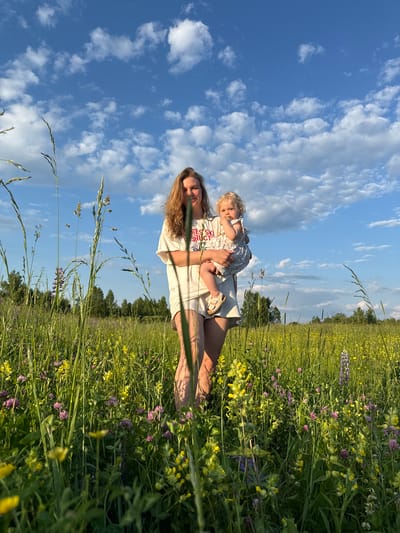 A woman and a young child are standing in a field of wildflowers under a blue sky with scattered clouds. The woman, with long blonde hair, is wearing a white t-shirt and light-colored shorts. She is holding the child in her arms. The child has blonde hair and is wearing a patterned dress. The field is dense with green grass, yellow and purple wildflowers, and tall stalks of grain. In the background, there is a line of trees. The image appears to have been taken in the vicinity of Колокша, Russia, a location known for its natural landscapes and agricultural fields.