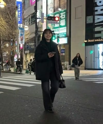 A nighttime urban scene depicts a bustling street in a commercial district, illuminated by numerous artificial lights and vibrant building signage. In the foreground, a young East Asian woman with dark, shoulder-length hair and bangs smiles directly towards the viewer while walking on a pedestrian crosswalk. She is dressed in a black puffer jacket, dark wide-leg pants, and dark flat shoes, holding a dark-colored smartphone in her left hand and a black handbag in her right. Overhead, a traffic light displays a red signal.

Buildings line both sides of the street, featuring multiple illuminated signs in Japanese. Prominent signs include a tall blue vertical sign on the left displaying "H&R" (エイチ・アンド・アール), and a large green rectangular sign with "石場" (Ishiba) and "職安ショップ" (Shokuan Shop – Employment Security Shop). A dark building on the right distinctly features the text "CROSS SHINJUKU" on its facade.

Further back on the right side, another young East Asian woman walks on the sidewalk, wearing a dark jacket over a light top and light pants, carrying a light-colored shoulder bag. On the far left, a masked individual cycles on a bicycle equipped with a green delivery bag. Other indistinct figures are visible on the sidewalks in the background. The street is asphalt with white parallel lines marking the crosswalk. Leafless trees are visible on the left, suggesting a cooler season.