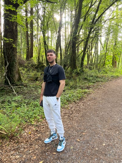 The image shows a man standing on a trail in a forest near Garraunbaun, Ireland. The man is wearing a black t-shirt, white sweatpants with a Nike logo, and light blue and white Nike high-top sneakers. He is facing the camera with his right hand on his hip. The trail is gravel and dirt. The forest consists of trees with green leaves and moss-covered trunks. The background is illuminated by sunlight filtering through the trees.