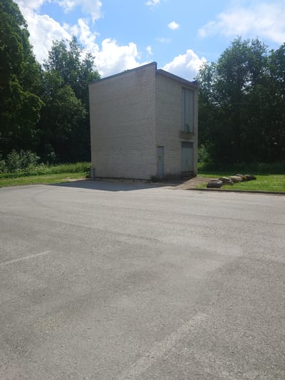 This image depicts a rectangular, light-colored brick building situated in an outdoor environment, likely a park or undeveloped area, with a paved surface in the foreground. The building features a simple design with a flat roof and two large, vertical windows or doors on its right side. A smaller, single door is visible on the left side of the building. The structure appears to be an isolated utility building or a small shed.
To the right of the building, a grassy area is visible, interspersed with several large, dark-colored rocks arranged in a line. Behind the building and to the left, a dense line of trees forms a natural backdrop. The sky above is blue with scattered white clouds, indicating fair weather. The paved area in the foreground suggests it could be a parking lot, driveway, or pathway adjacent to the building.
The location is identified as being in Alavere, Estonia. The scene is static, with no people or apparent activity. The overall impression is one of a functional, utilitarian structure set within a natural, somewhat unkempt, landscape. The image is taken from a low-angle perspective, looking slightly upwards towards the building and the sky.