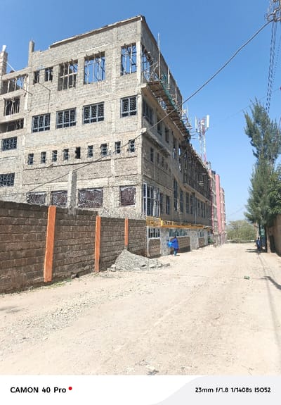 The image depicts a multi-story building, appearing to be five to six floors high, constructed from concrete blocks in Oloosirkon/Sholinke ward, Kenya. The building is in various stages of construction, with the upper floors showing exposed concrete and rebar, open window frames, and scaffolding partially covered with green netting on the right side. Lower floors feature installed windows with dark frames and security bars, while the ground floor has white-framed windows and doors. A yellow sign above the ground floor entrance reads "JESUS VOICE MINISTRIES." To the left, a stone wall with orange vertical pillars and barbed wire on top runs alongside an unpaved, dusty dirt road. A pile of grey gravel and some loose stones are present on the road near the building's base. One individual in a blue garment stands near the "JESUS VOICE MINISTRIES" entrance, facing away from the camera. Further down the road on the right, another person in a dark shirt walks away from the camera, partially obscured by a tree. In the background, a tall, white and red cell tower with antennae is visible, alongside numerous electrical or communication wires traversing the clear blue sky. A pink/red multi-story building and other structures are visible further down the road to the right, bordered by trees.