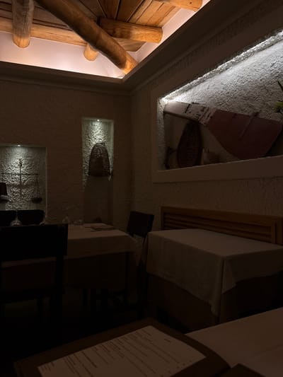 The image shows the interior of the restaurant "Il Rifugio del Contadino" in Agropoli, Italy. The walls have a textured, stucco-like finish, and recessed niches are illuminated with warm light, displaying decorative items like a basket and a vintage scale. The left side features a wooden structure, possibly part of the building's rustic design. Tables are set with white linens, and a menu is visible in the foreground.