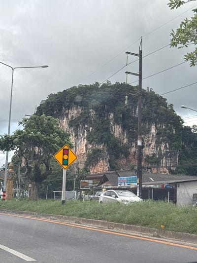 A traffic light sign, depicting red, yellow, and green lights, is prominently displayed on a yellow diamond-shaped sign. The sign is positioned to the left of a paved road with a yellow line marking its edge. To the left of the road, a steep, lush green mountain face rises dramatically. A white van is parked in front of a building that appears to be a shop, partially obscured by overgrown grass and vegetation. Utility poles and overhead wires are visible, suggesting an urban or semi-urban environment. The sky is overcast, indicating a cloudy or possibly rainy day. The overall scene suggests a roadside view in a tropical or subtropical region, given the presence of the mountain and dense vegetation. No people are visible in the image. The time of day is unclear due to the overcast conditions. Text is present on a blue sign attached to a building, but it is too blurry to read.