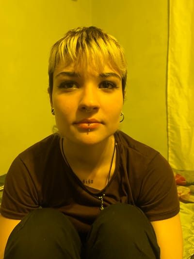 A rotated, indoor photograph depicts a fair-skinned person with blonde and dark hair, wearing a dark brown short-sleeved t-shirt and dark pants. The person is angled towards the camera. They have piercings in their nose and lower lip, and visible tattoos on their neck. The background includes a yellow wall and a bed with a patterned blanket. The location in Campana, Argentina cannot be determined from the image.