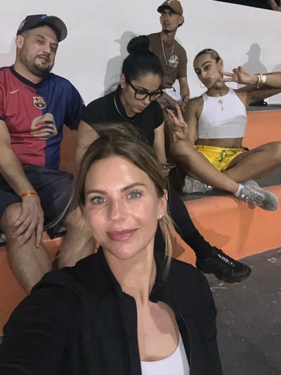 The image depicts six individuals seated on orange-colored bleachers or steps against a plain white wall, likely in San Pedro Town, Belize, as indicated by a visible wristband. The setting suggests an indoor or covered outdoor venue with artificial lighting, evidenced by shadows cast on the wall.

In the immediate foreground, a fair-skinned female with blonde, shoulder-length hair and blue eyes is taking a selfie, looking directly at the camera with a slight smile. She is wearing a black zipped jacket over a white top and has small silver stud earrings.

To her left, a male with a beard and mustache, wearing a reversed dark blue baseball cap, is seated. He is wearing a red and blue split FC Barcelona jersey with the club crest and a stylized blue, white, and red hand design on the lower portion. He wears dark shorts, and an orange wristband on his right wrist clearly displays "San Pedro Town." He is also looking towards the camera with a faint smile.

Behind the foreground woman and next to the Barcelona fan, a female with dark hair pulled into a high bun is visible. She wears black-framed glasses and a black long-sleeved top, looking downwards or away from the camera.

Further back and to the right of the central woman, a slender male wearing a brown baseball cap, a brown t-shirt with a white graphic, and a silver chain with pendants is seated. He has a light mustache and goatee and is looking towards the right side of the frame.

To the right of the central group, a slender female with light hair in a slick ponytail is seated. She wears a white crop top, bright yellow athletic shorts with a green design, and light gray athletic shoes with white socks. Her right wrist displays multiple bracelets, and she has several rings on her fingers. She is making a "peace" sign with both hands, looking towards the camera with a slight pout, and has visible hoop earrings.

All individuals are seated in close proximity, suggesting a shared activity or gathering.