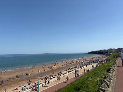 The image depicts a sunny day at Longsands Beach in Tynemouth, North Tyneside, United Kingdom. A wide, sandy beach stretches along the coastline, populated with numerous people sunbathing, swimming, and walking. The sea is calm and blue, with several distant ships visible on the horizon. Adjacent to the beach is a paved promenade, also populated with pedestrians. A stone wall separates the promenade from a grassy verge. In the background, buildings line the coastline. The sky is clear and blue, indicating a sunny day.
