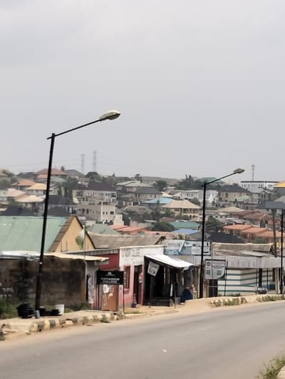 The image displays a daytime street scene in Otta, Nigeria, under an overcast, light grey sky. In the foreground, an asphalt road ascends slightly from the lower center, bordered on its right by a curb with black and yellow diagonal stripes. Two black lampposts, each with a white, oval-shaped luminaire, are positioned along the right side of the road; one is prominent on the left, and the other recedes towards the right. Immediately adjacent to the road on the right are several low-rise structures: a faded reddish-pink building with a brown door featuring a "TOTARAM COLLEGE" sign, followed by an open-front stall with an extending corrugated metal roof, and then a building with horizontal green and white striped siding. On the left side of the road, a low concrete wall is visible. The midground presents a dense urban sprawl of single and multi-story buildings with varied roof colors including green, orange, brown, and grey. A green and white flag is visible on a building with a green roof on the left. The background extends into a residential area covering a hillside, characterized by numerous houses with diverse roof types. In the far distance, several metal lattice electricity transmission towers and a telecommunications mast are visible against the sky. No individuals, vehicles, or dynamic actions are depicted.