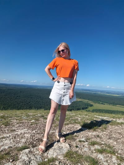 A woman with blonde hair, wearing sunglasses, an orange crop top, and a white denim skirt, stands on a rocky hill. She is posing with one hand on her hip. The background shows a vast green landscape with a forest and fields under a clear blue sky. The location is the city of Shikhan, Russia.