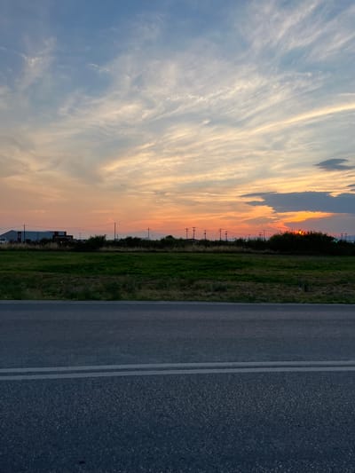 The image captures a sunset over a rural landscape in Chaidefto, Greece. The sky is filled with wispy clouds painted in hues of orange, pink, and yellow, indicative of the golden hour. Silhouetted against this vibrant sky are utility poles and a line of trees, suggesting a flat terrain. To the right of the frame, a paved road runs parallel to a grassy field. The road is marked with two white lines, denoting a lane divider. The overall scene evokes a sense of tranquility and the beauty of nature as day transitions into night. No people are visible in the image. A building with the text "MARINE CENTER" is partially visible in the distance.