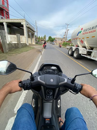 The image captures a first-person perspective from a motorcycle in Cottrueng, Indonesia. The rider, wearing blue jeans and flip-flops, is navigating a road alongside a large white tanker truck. The truck is branded with "PERTAMINA PATRA NIAGA" in red and blue lettering. The road is paved and has a yellow line dividing the lanes. On the left side of the road, there are houses and a building under construction. Power lines are visible overhead, and palm trees suggest a tropical climate. The sky is overcast, indicating potentially cloudy weather. The overall scene depicts everyday life and transportation in a semi-urban or rural Indonesian setting. The rider's hands are on the handlebars of the motorcycle, suggesting the act of riding. The image is taken during the daytime, as indicated by the lighting.